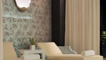 Quarto de hotel moderno com duas sofás, um detalhe decorativo de conchas na parede, um vaso de plantas e uma grande janela com cortinas.