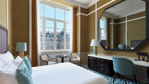 A hotel room with a large bed, a desk, a chair, a dresser, and a large window offering a city view. - The One Monumental Palace (Porto)