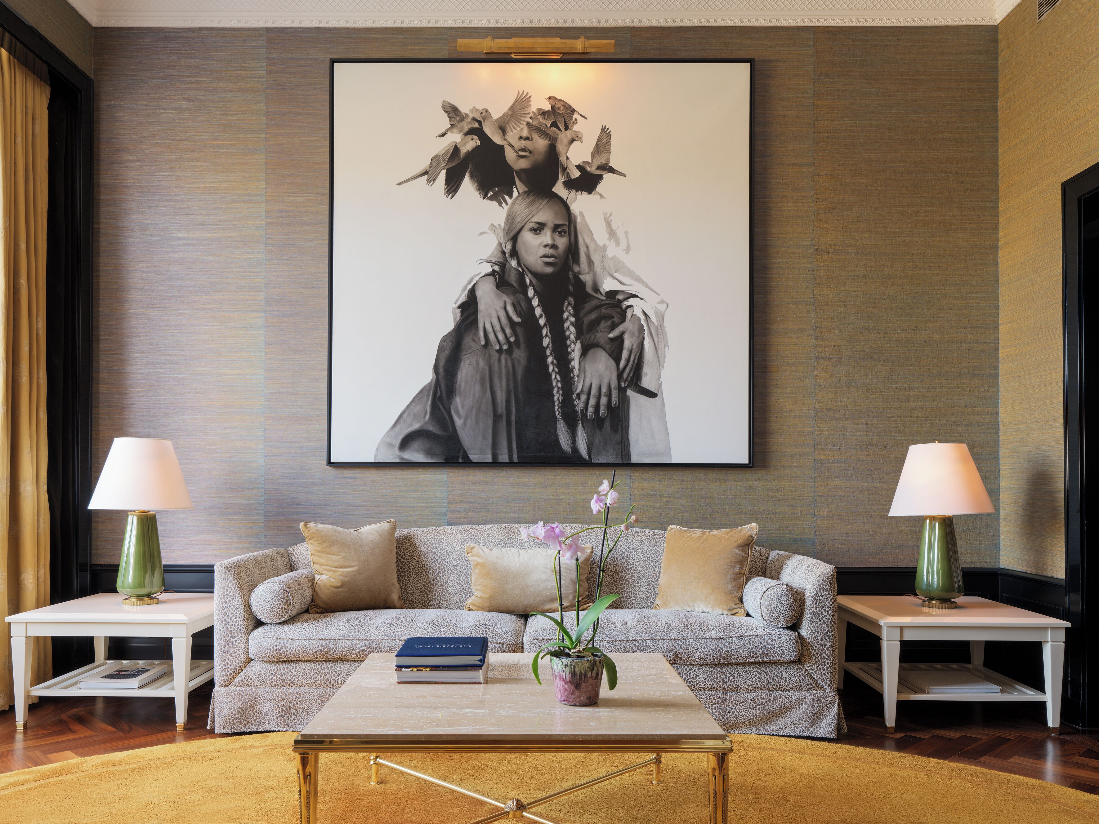 Een woonkamer met een groot kunstwerk in een lijst aan de muur, een bank met gekleurde kussens, een salontafel met boeken en een plant, en twee bijzettafels met lampen.