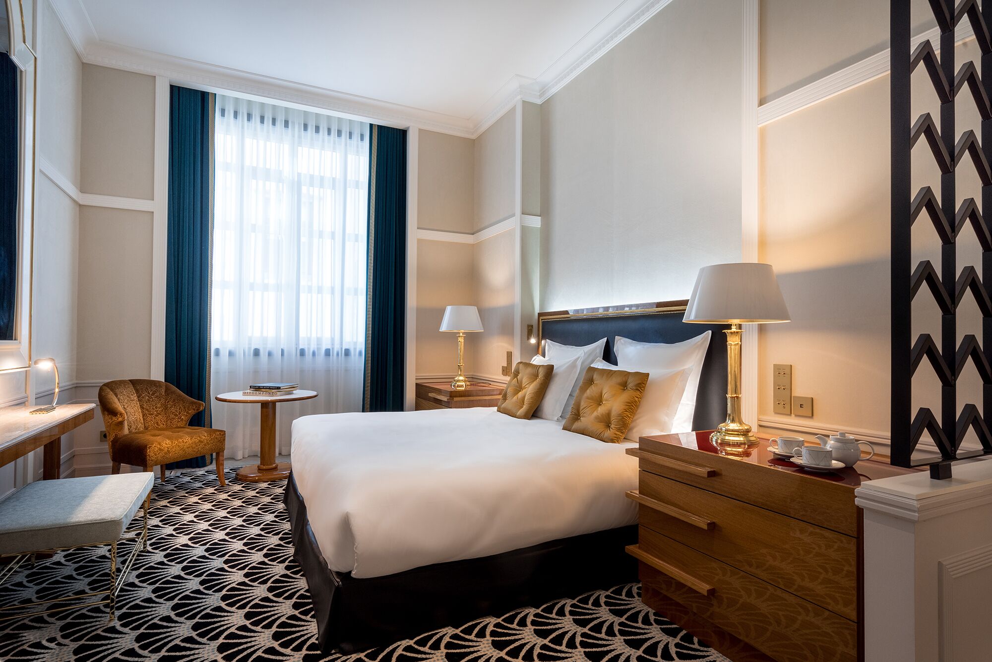 A hotel room with a large bed, bedside tables, a chair, a desk, and a patterned rug.