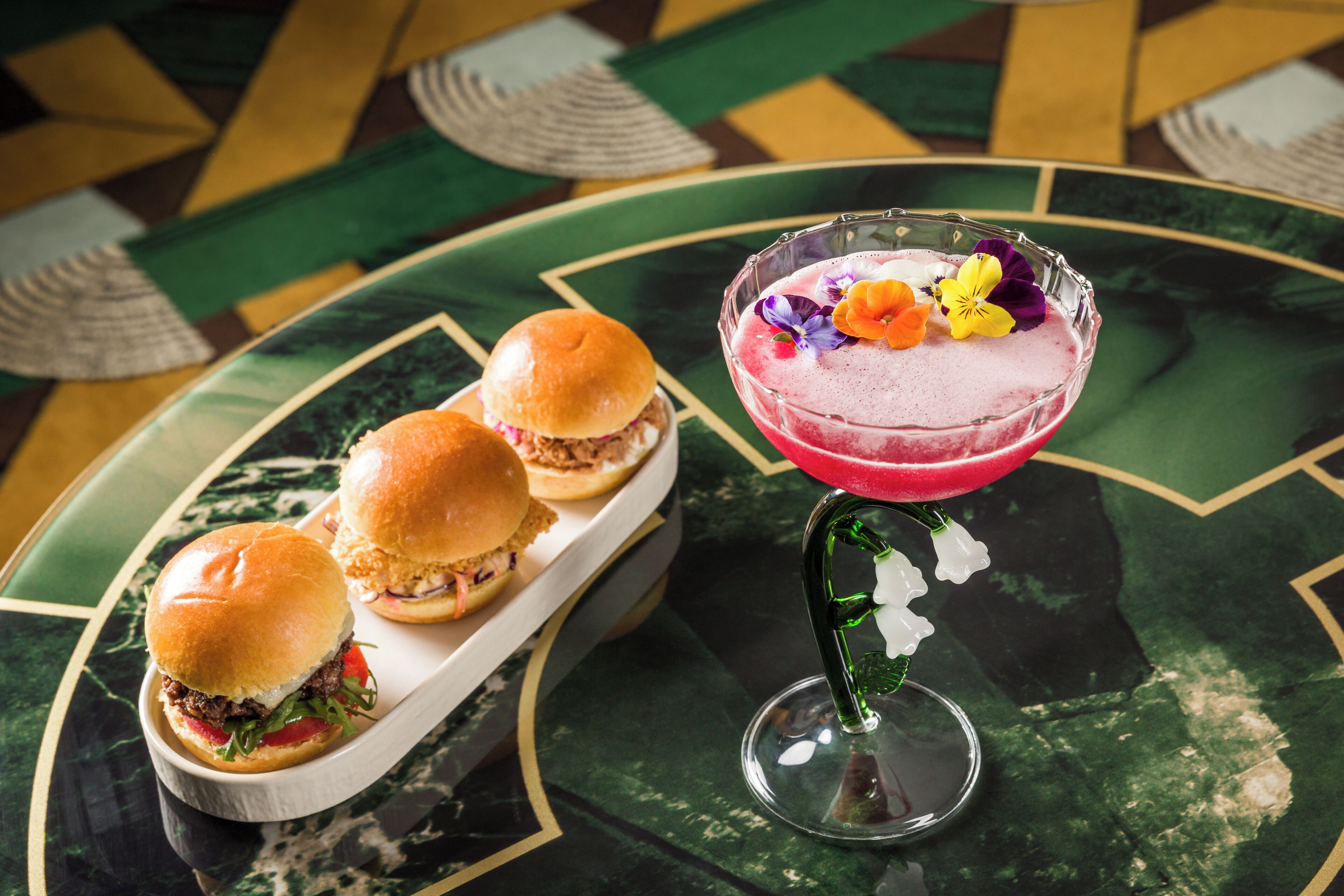 Une table avec trois hamburgers et un cocktail décoré de fleurs.