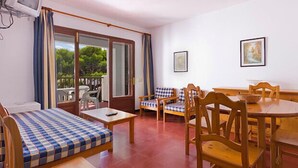 A room with a checkered sofa, a wooden dining table with chairs, a small table with a bowl, and a view through a glass door to an outdoor area with a chair and a table. - Apartamentos Sol Y Mar Menorca (Ciutadella de Menorca)