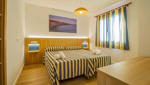 A hotel room with a double bed, a wooden headboard, a bedside table, a nightstand, a desk, a lamp, a window with curtains, and a painting on the wall. - Apartamentos Sol Y Mar Menorca (Ciutadella de Menorca)