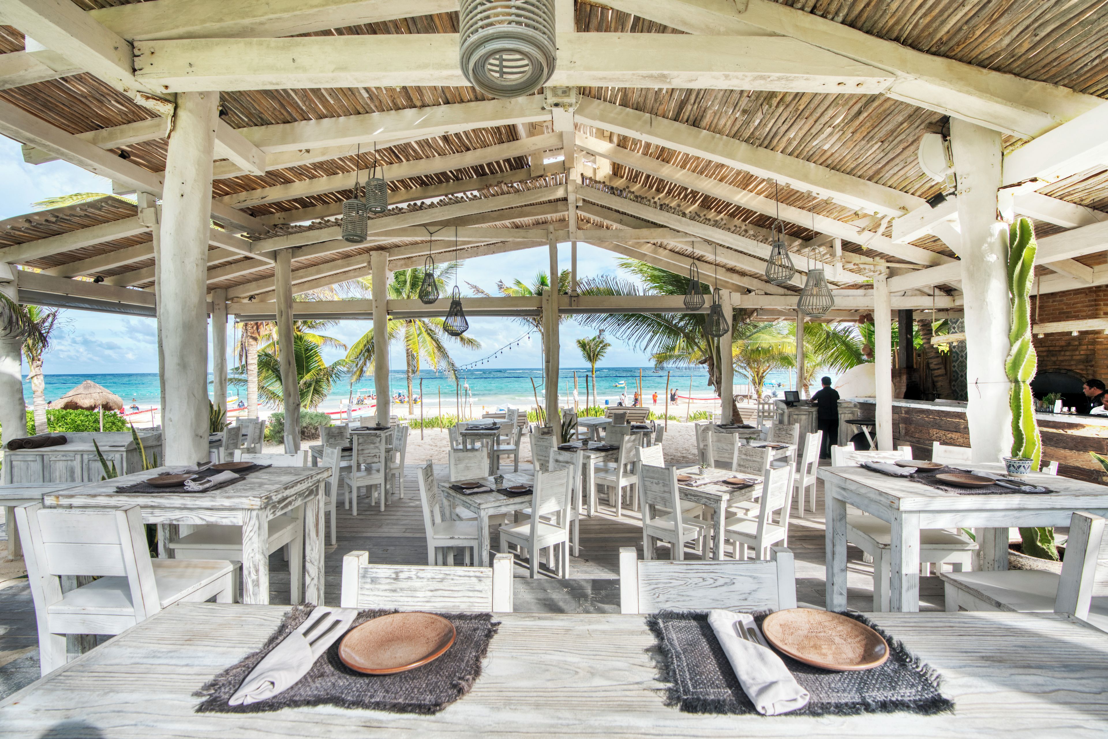 An outdoor dining area with white tables and chairs, a thatched roof, and a view of the beach and ocean.