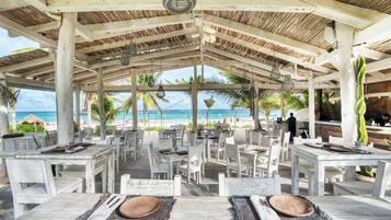 An outdoor dining area with white tables and chairs, a thatched roof, and a view of the beach and ocean.