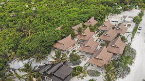 Vue aérienne d’un complexe hôtelier avec des villas aux toits de chaume, une plage de sable blanc et une végétation luxuriante.