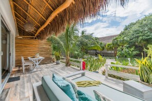 Un patio exterior con techo de paja, zona de estar con cojines, mesa y sillas, y diversas plantas.
