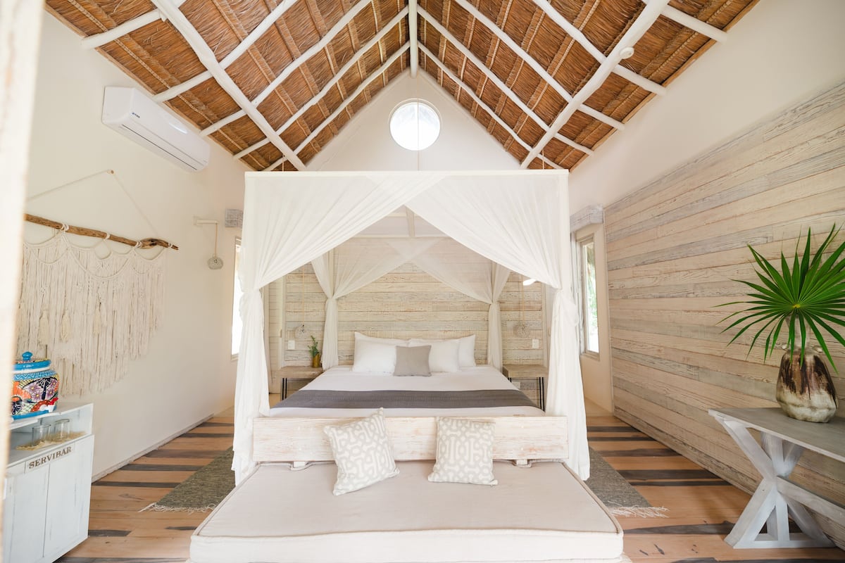 A room with a bed under a canopy, a wooden wall, a plant, and a table.