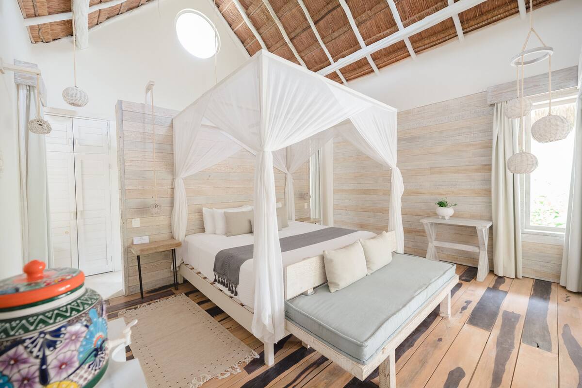 A bedroom with a four-poster bed, a small bedside table, a nightstand, a decorative vase, and a window with curtains.