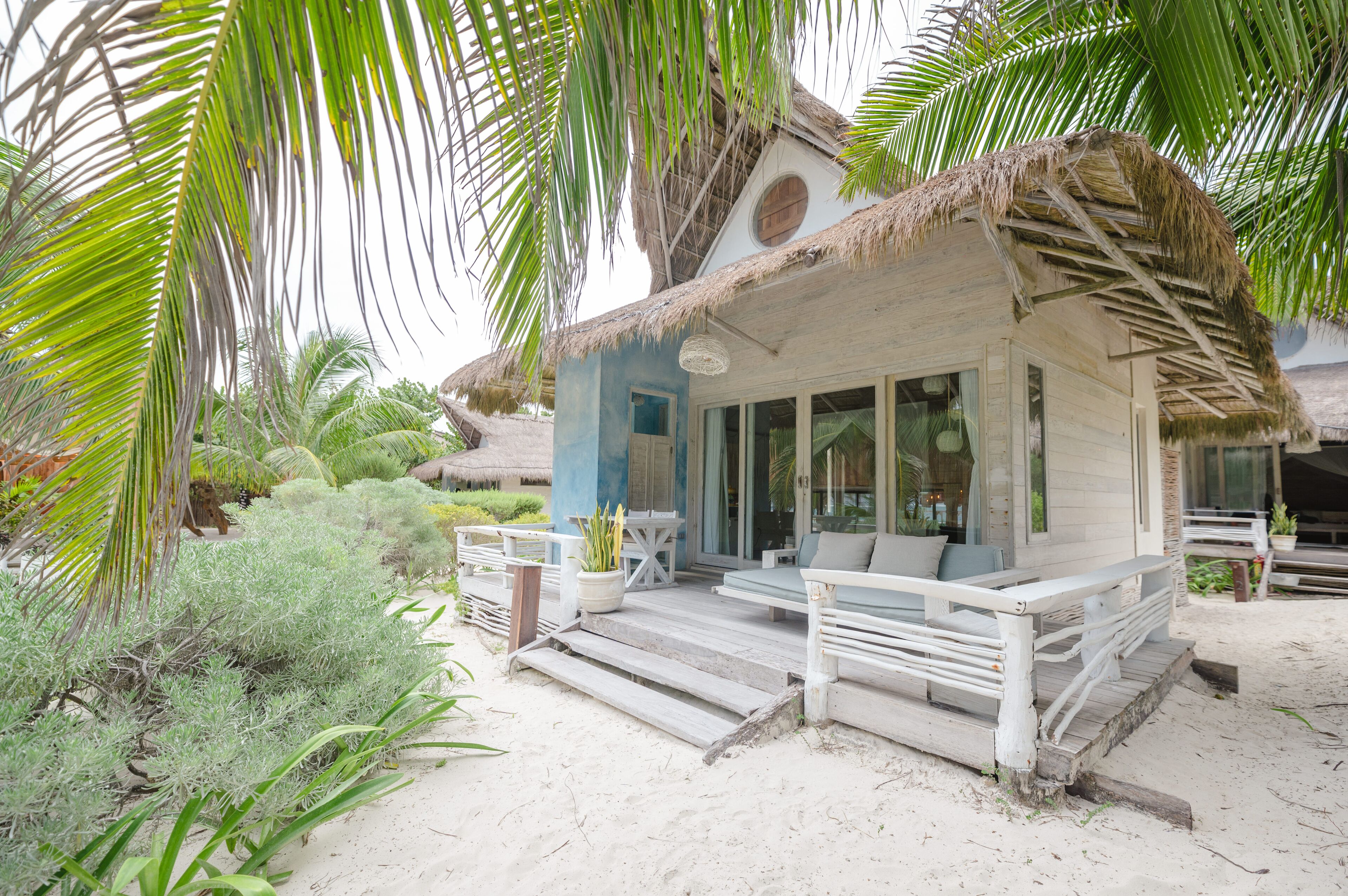 A beach house with a thatched roof, a porch with white railings, and a sandy beach area.
