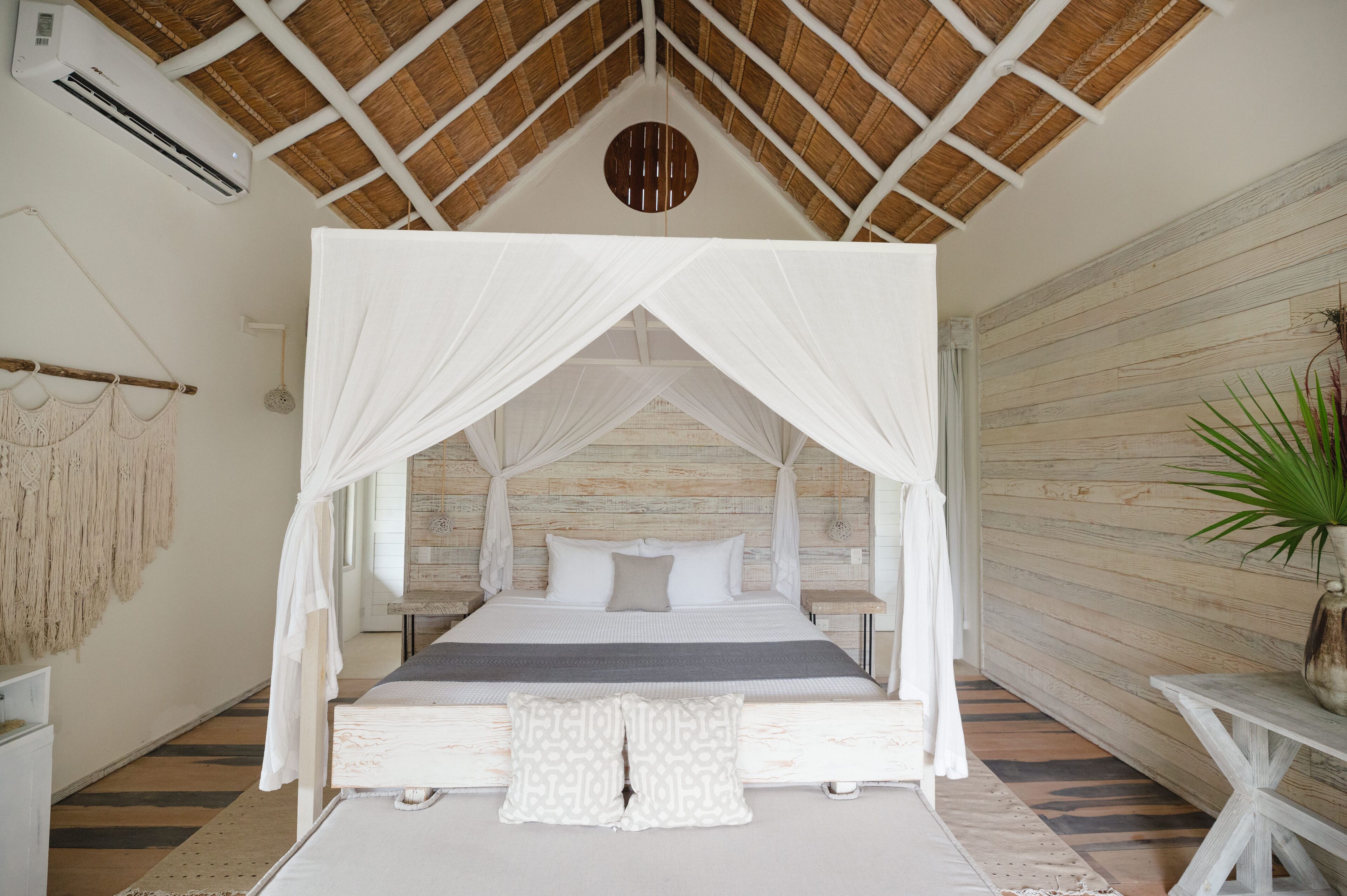 A room with a bed under a canopy, a wooden wall, and a plant on a table.