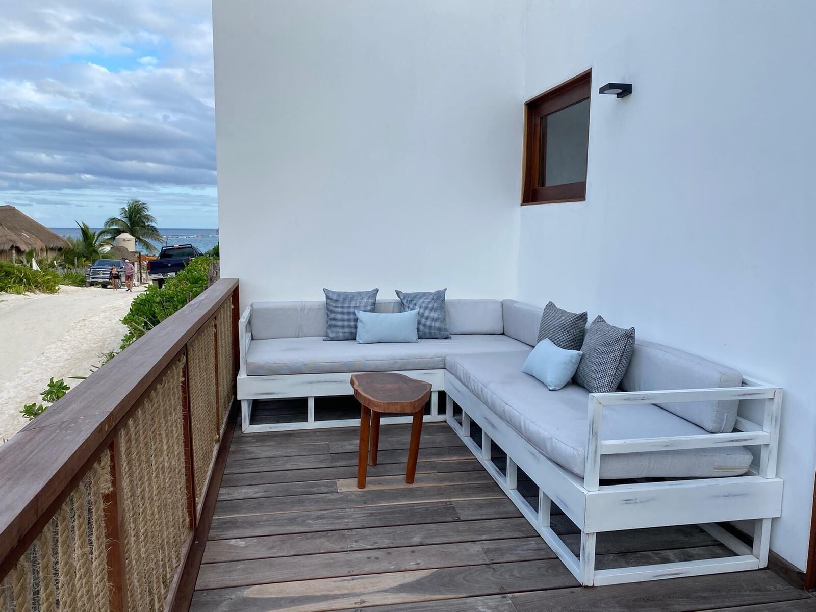 Un espacio al aire libre con asientos blancos, una mesa de madera, un sofá acolchado con varios cojines y una barandilla de madera.