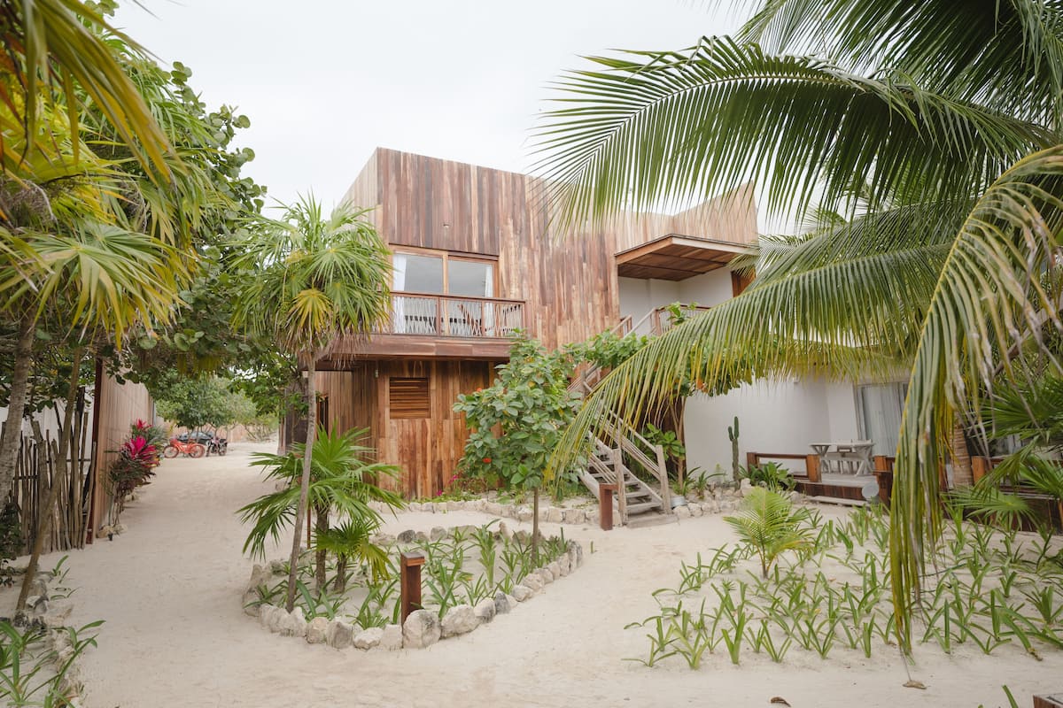A modern building with a wooden facade, a balcony, and a sandy courtyard surrounded by tropical plants.