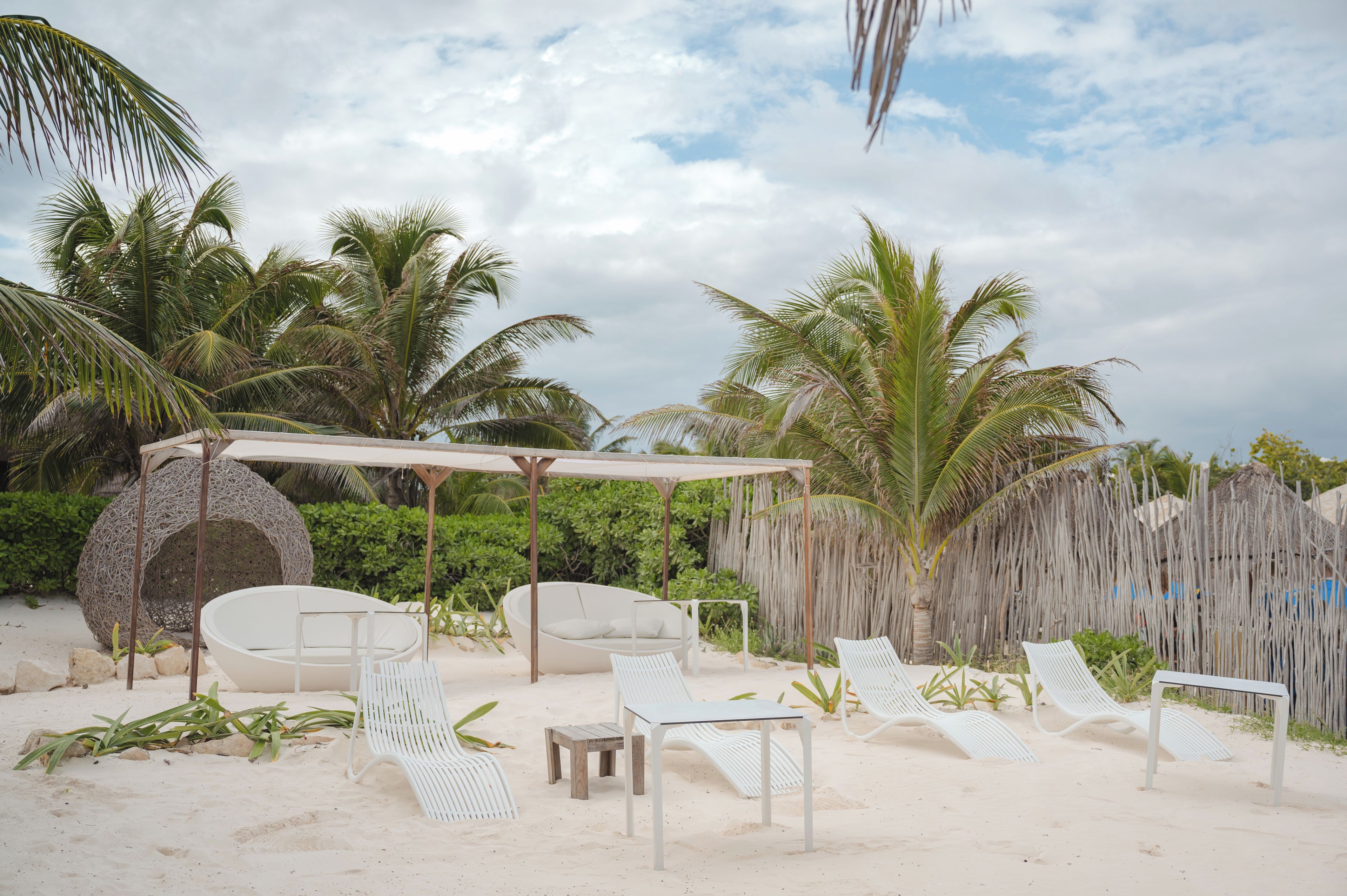 A beachside lounge with white lounge chairs and a covered seating area, surrounded by palm trees and a thatched wall.