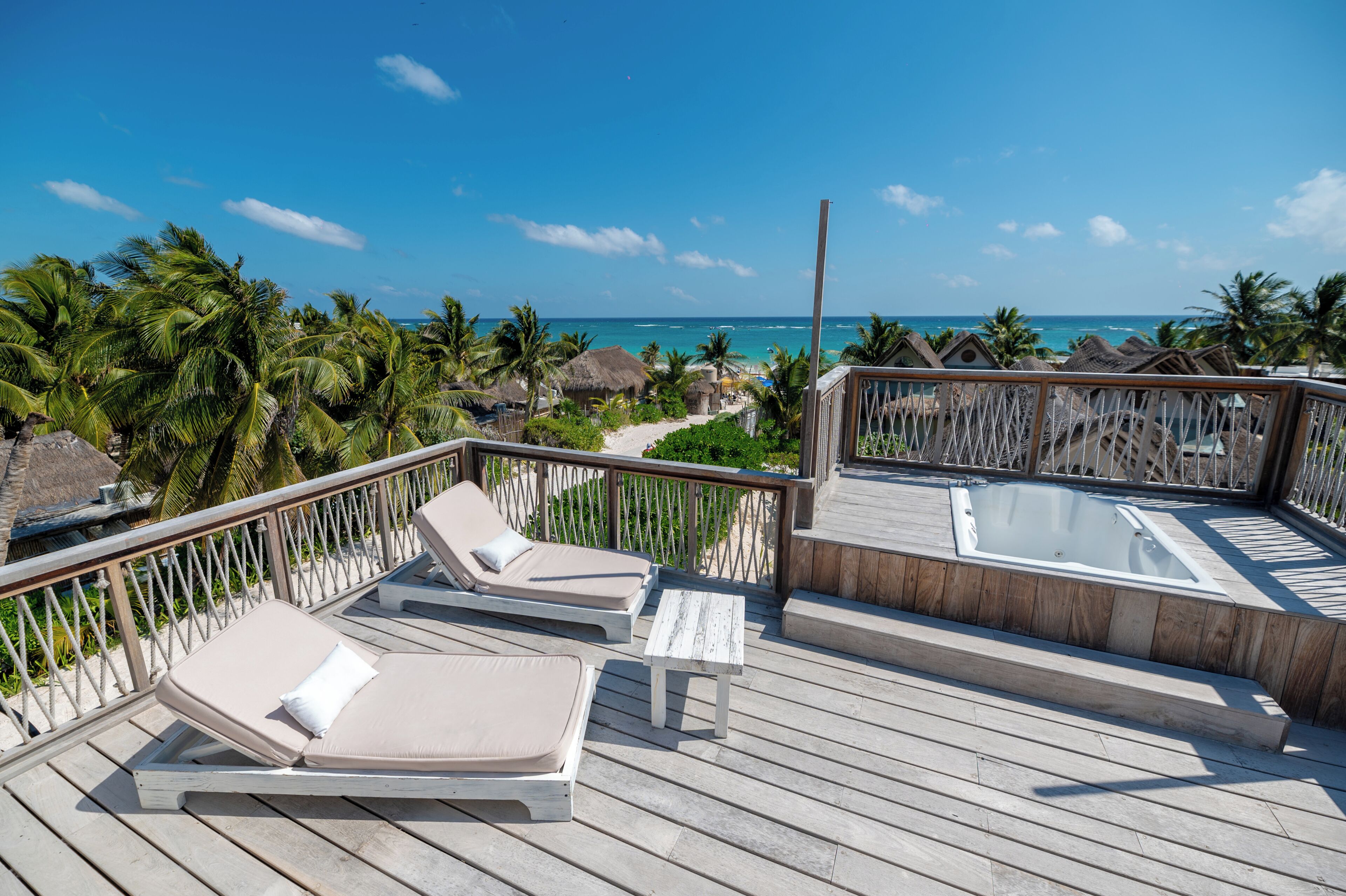 Um deck de madeira com cadeiras de descanso e uma banheira de hidromassagem, com vista para uma praia com palmeiras e palafitas.