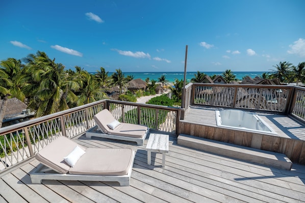 A wooden deck with lounge chairs and a hot tub, overlooking a beach with palm trees and thatched-roof huts.