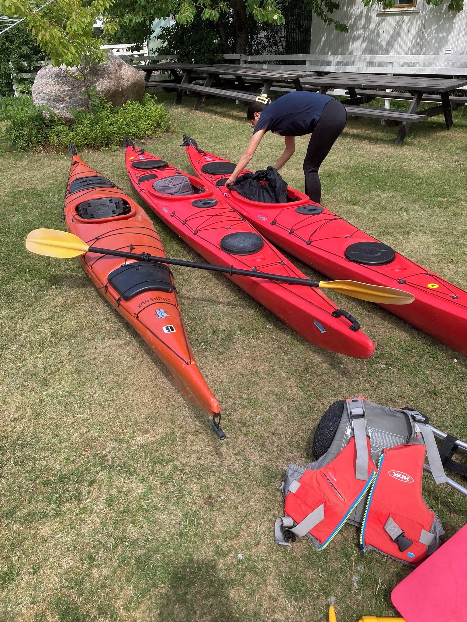 Two red kayaks, a paddle, and a red life jacket on a grassy area with picnic tables in the background.