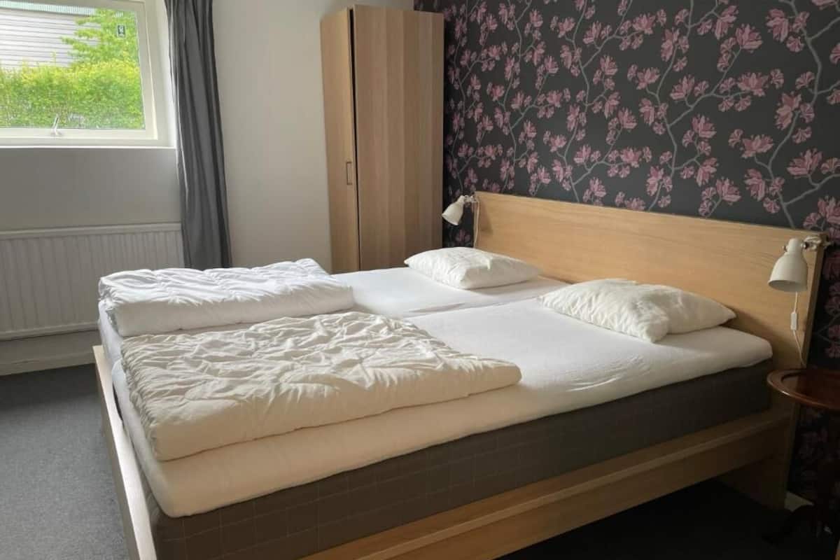 Una cama bien hecha con cabecera de madera, una mesita de noche con lámpara y una ventana con cortinas.