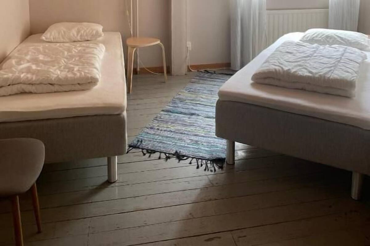 Un dormitorio con dos camas, techo de madera, ventana con cortinas y un taburete pequeño.