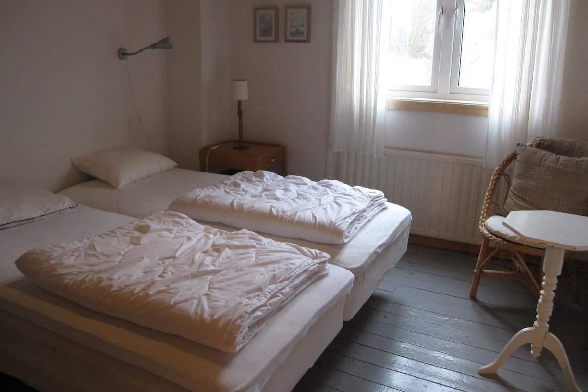 Habitación de hotel con dos camas, una mesita, una silla, una lámpara y una ventana con cortinas.