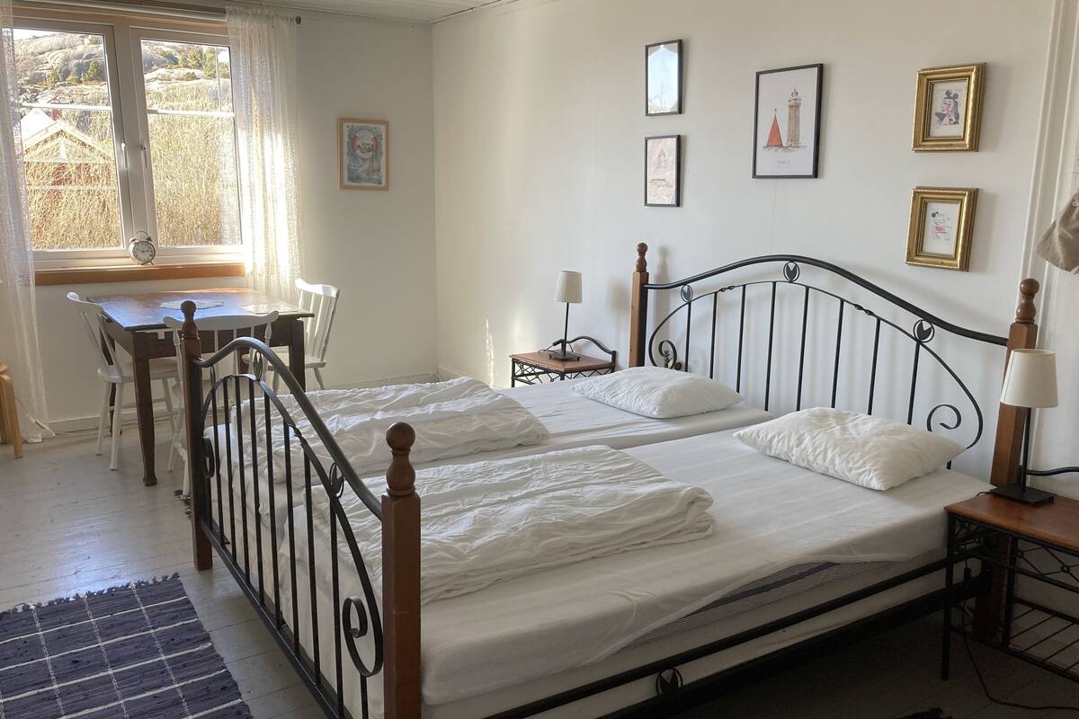 Cama doble con ropa de cama blanca, cabecera de madera y mesita de noche con lámpara. También se observa un escritorio con silla y una ventana con cortinas.