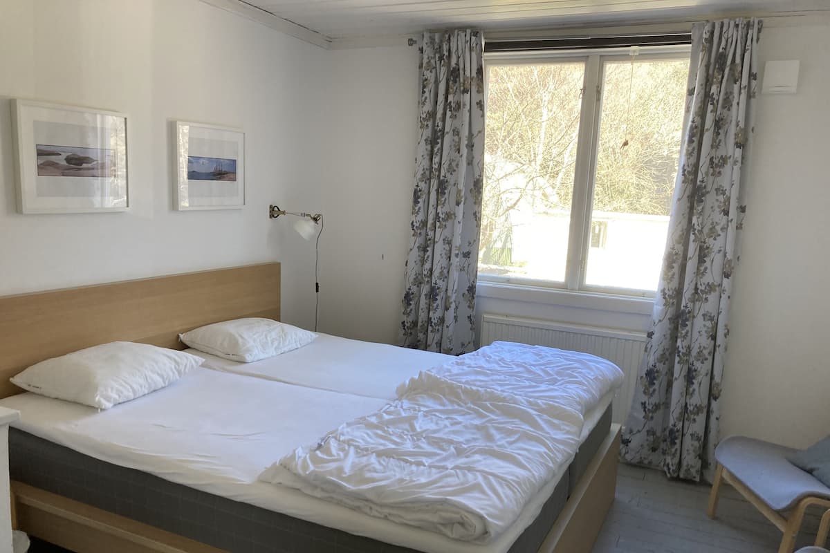 Una cama doble con sábanas blancas, un cabecero de madera, una lámpara de noche y dos cuadros en la pared.