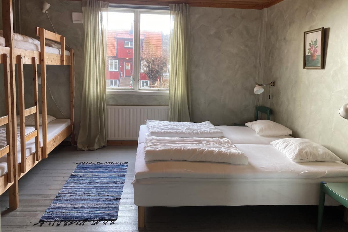 Una habitación con una cama litera, una cama individual, una ventana con cortinas, un radiador, una lámpara y un cuadro en la pared.