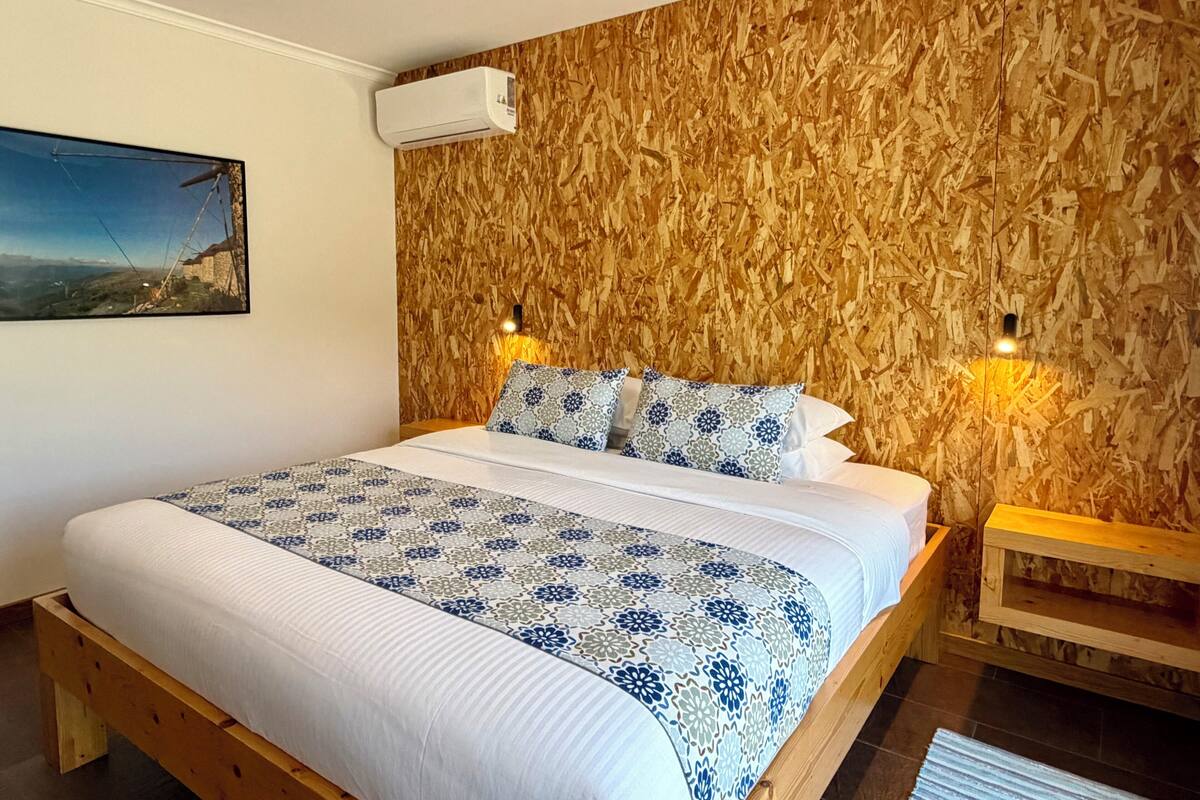 Un dormitorio con cabecera de madera, cama con ropa de cama estampada, aire acondicionado de pared y mesita de noche con lámpara.