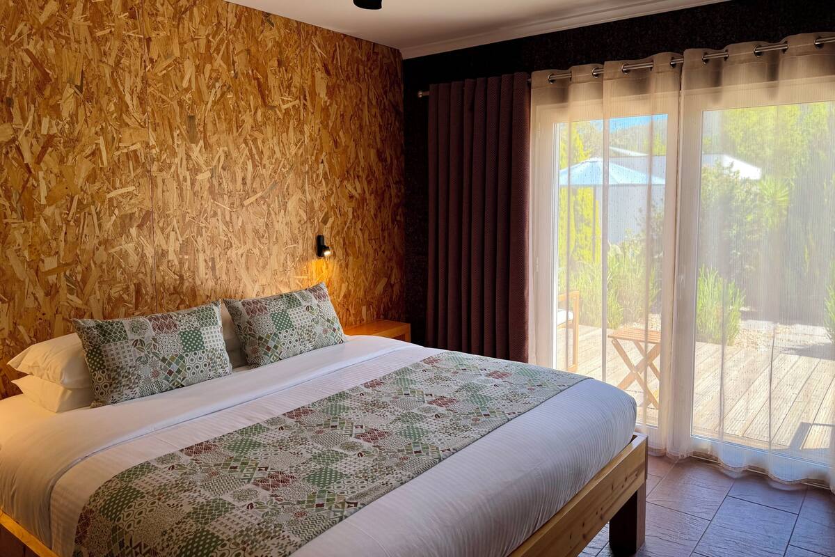 Un dormitorio con pared de madera, cama con ropa de cama estampada, ventilador de techo y vistas al exterior a través de una ventana con cortinas.
