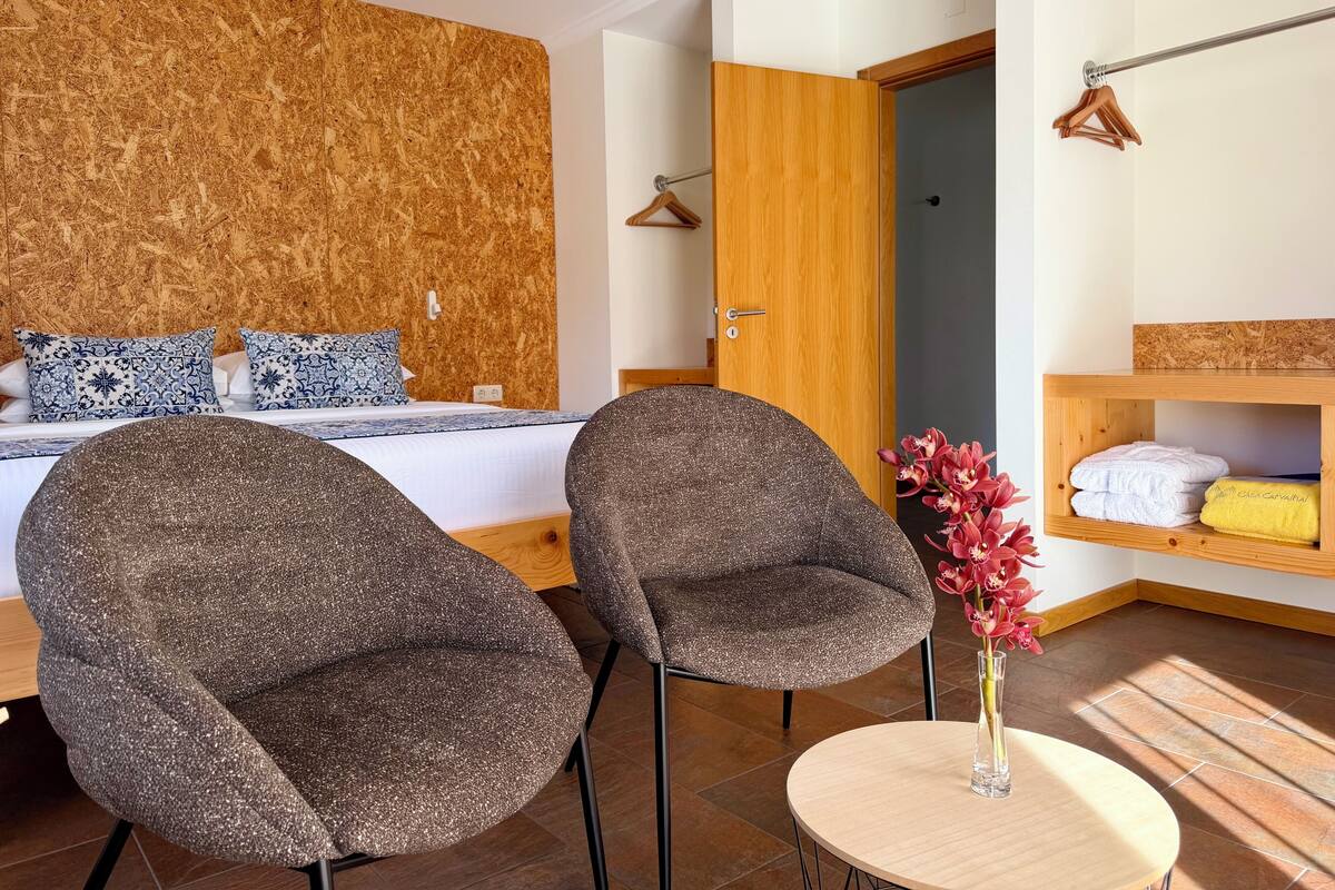 Un dormitorio con una cama, dos sillas, una mesita con un florero y un estante de madera con toallas.
