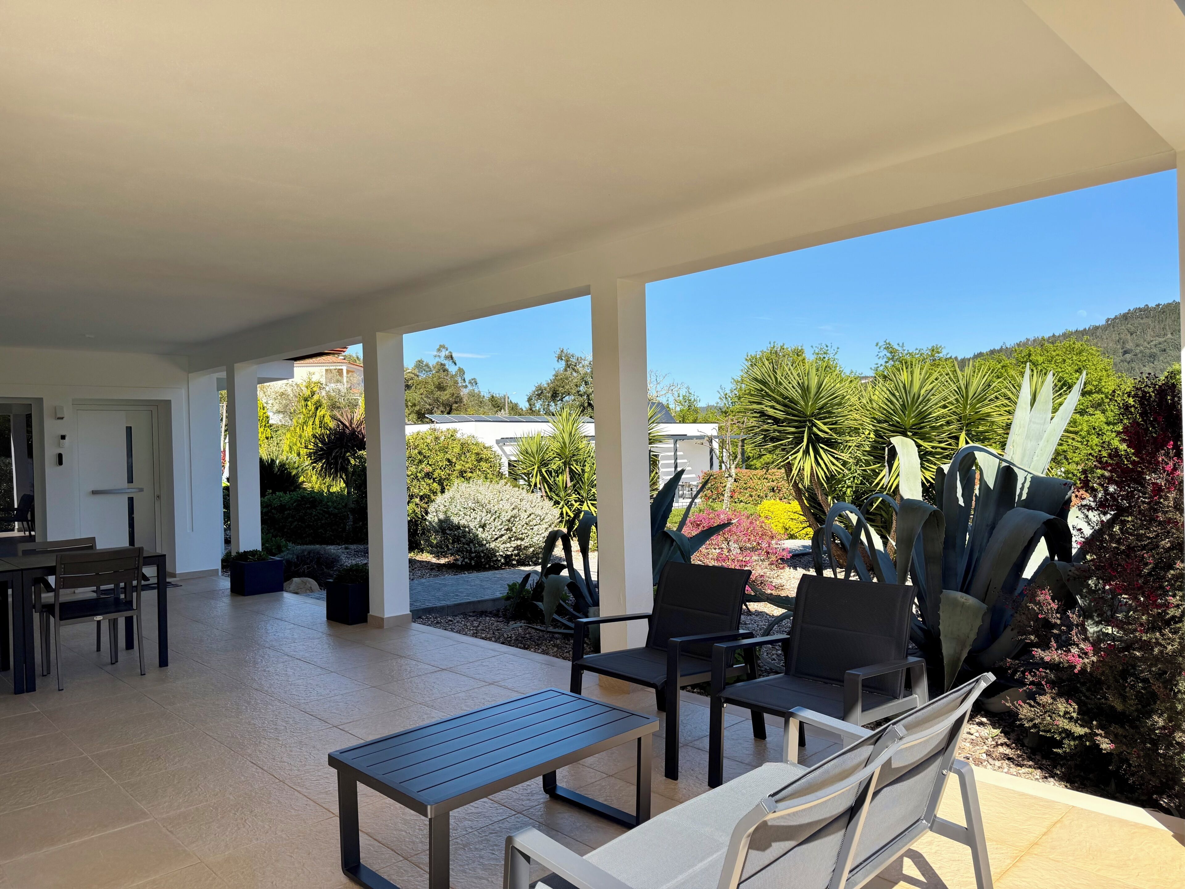 A covered patio with a dining table and chairs, surrounded by various plants and a view of the landscape.