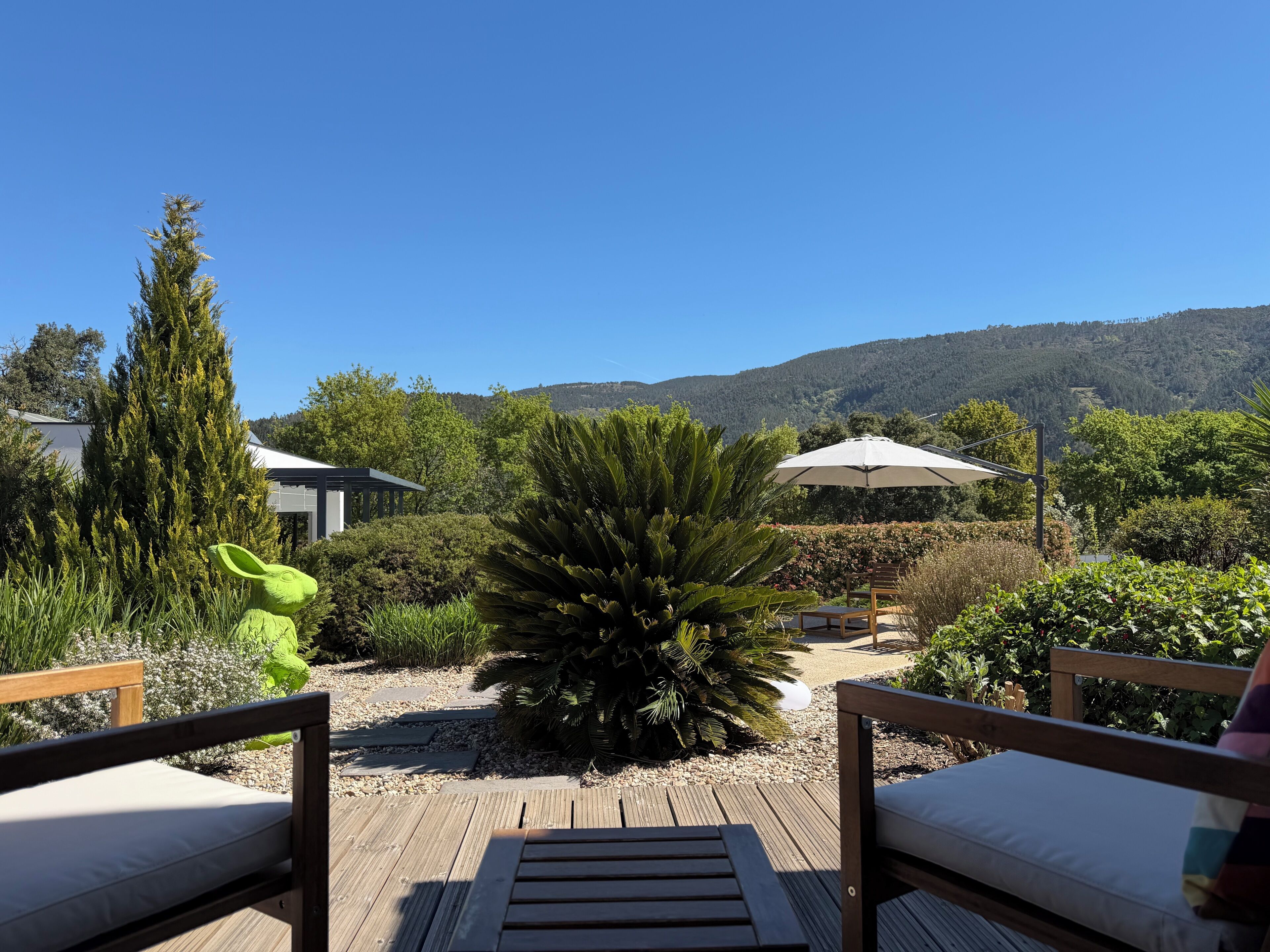 Een houten terras met twee stoelen, een tuin met diverse planten, een witte parasol en een berg op de achtergrond.