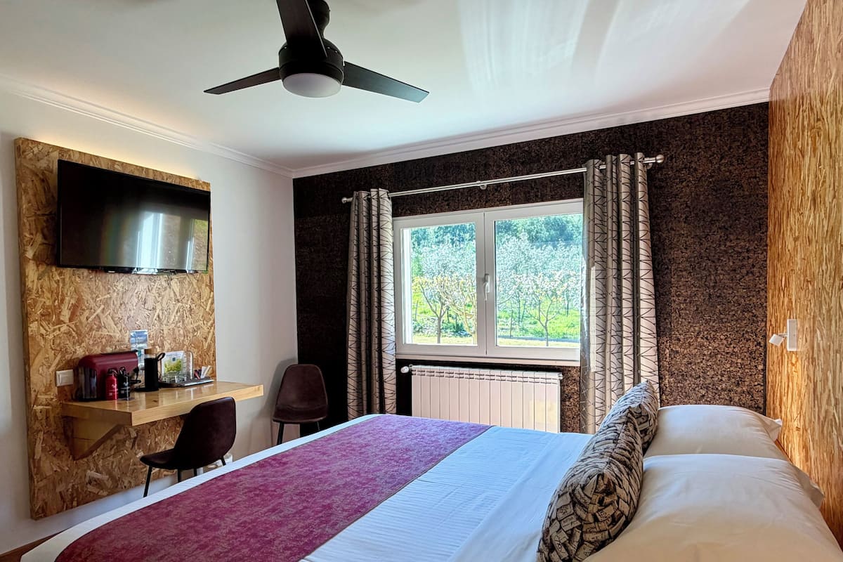 Habitación de hotel con cama, televisor montado en una pared de madera, ventana con cortinas y ventilador de techo.