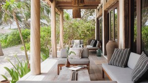 A covered outdoor seating area with a sofa, chairs, and a table, surrounded by greenery and a sandy path.