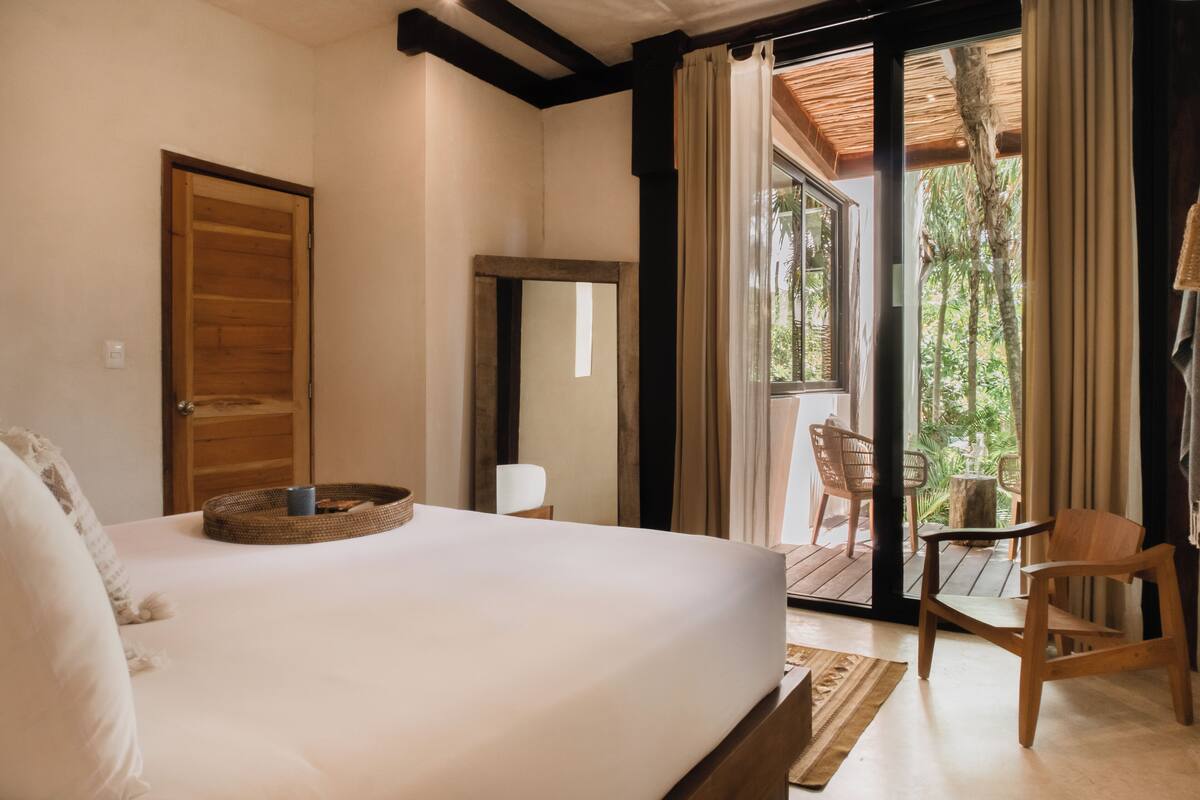 Une chambre avec un grand lit, une porte en bois, un miroir et une vue sur une terrasse extérieure avec des chaises.
