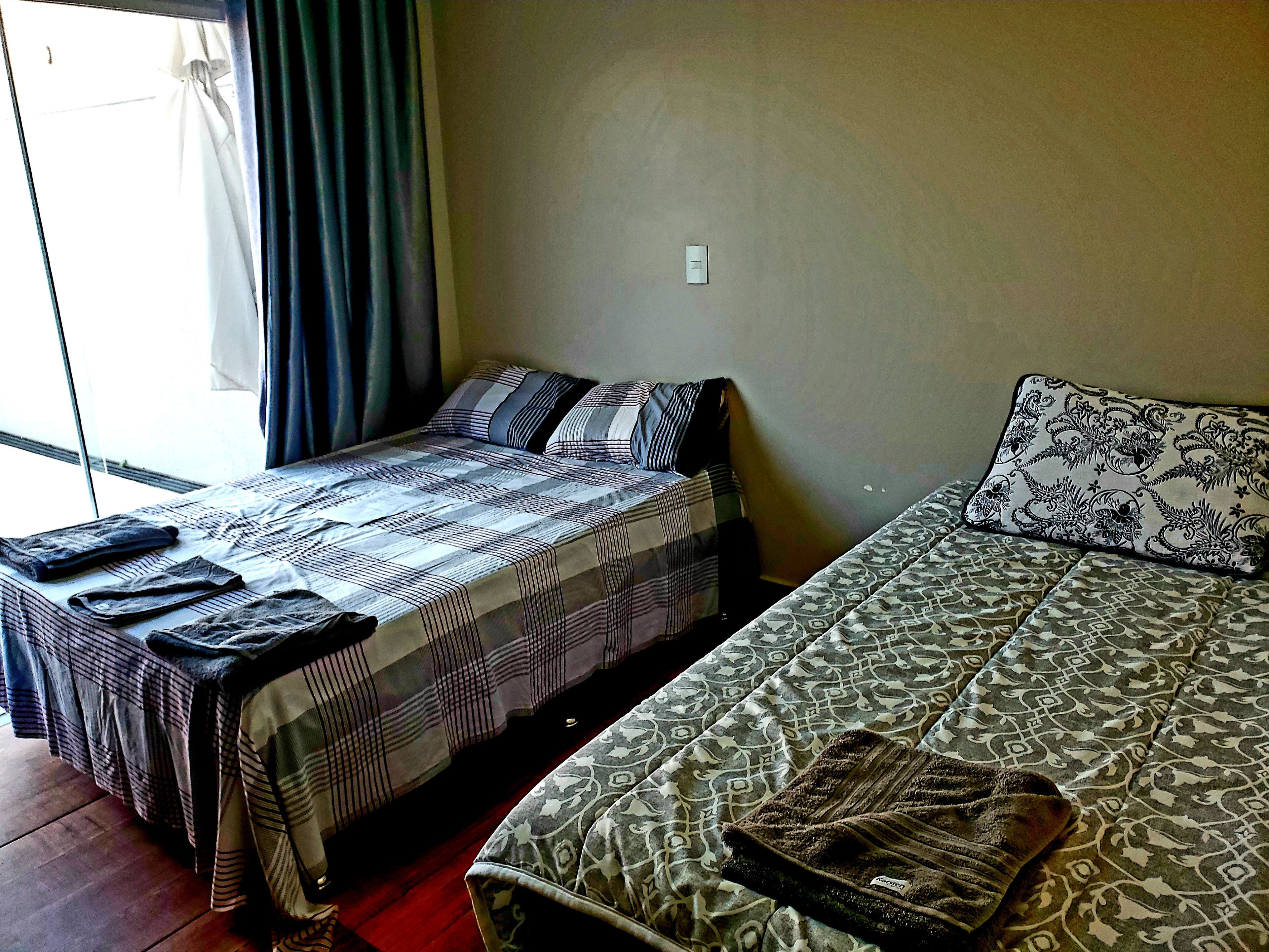 Une chambre avec deux lits, l’un recouvert d’une couvre-lit à rayures et l’autre d’une couvre-lit à motifs. Il y a une fenêtre avec des rideaux, un interrupteur au mur et un petit tapis sur le sol.