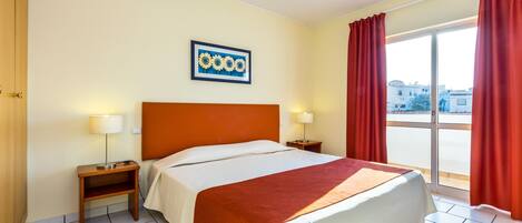 Habitación de hotel con cama, dos mesitas de noche, cortinas rojas y vista a la ciudad.