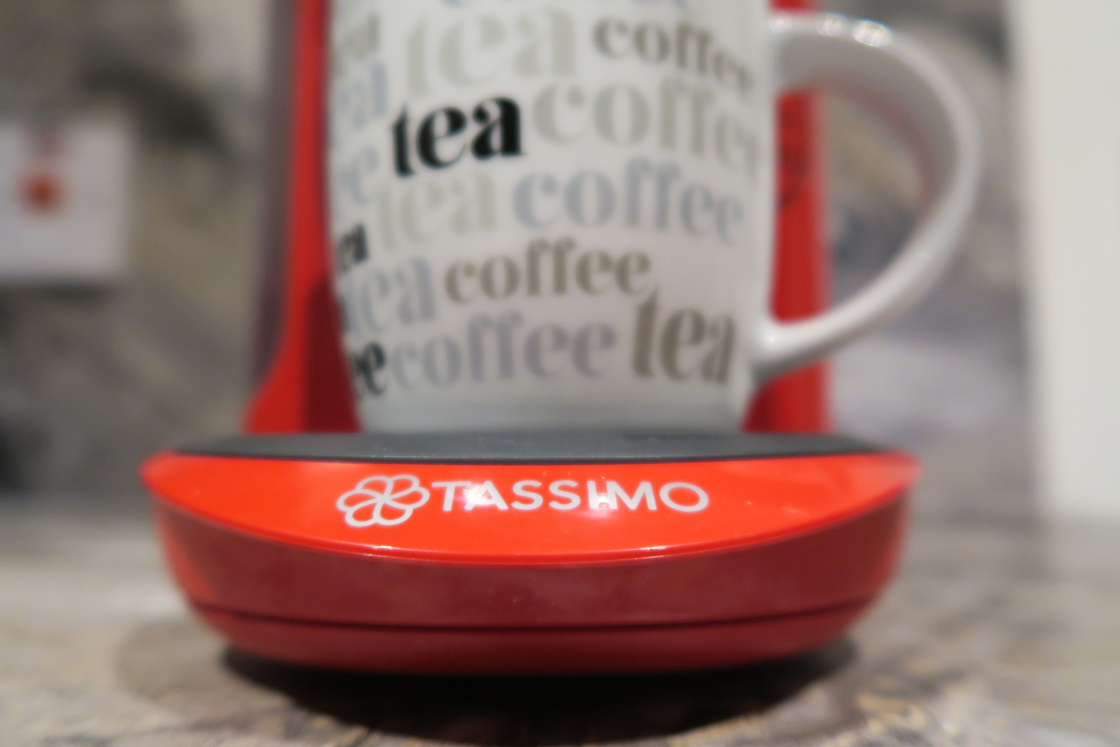 Une capsule Tassimo rouge est posée sur un comptoir de cuisine, avec une tasse blanche floue en arrière-plan.