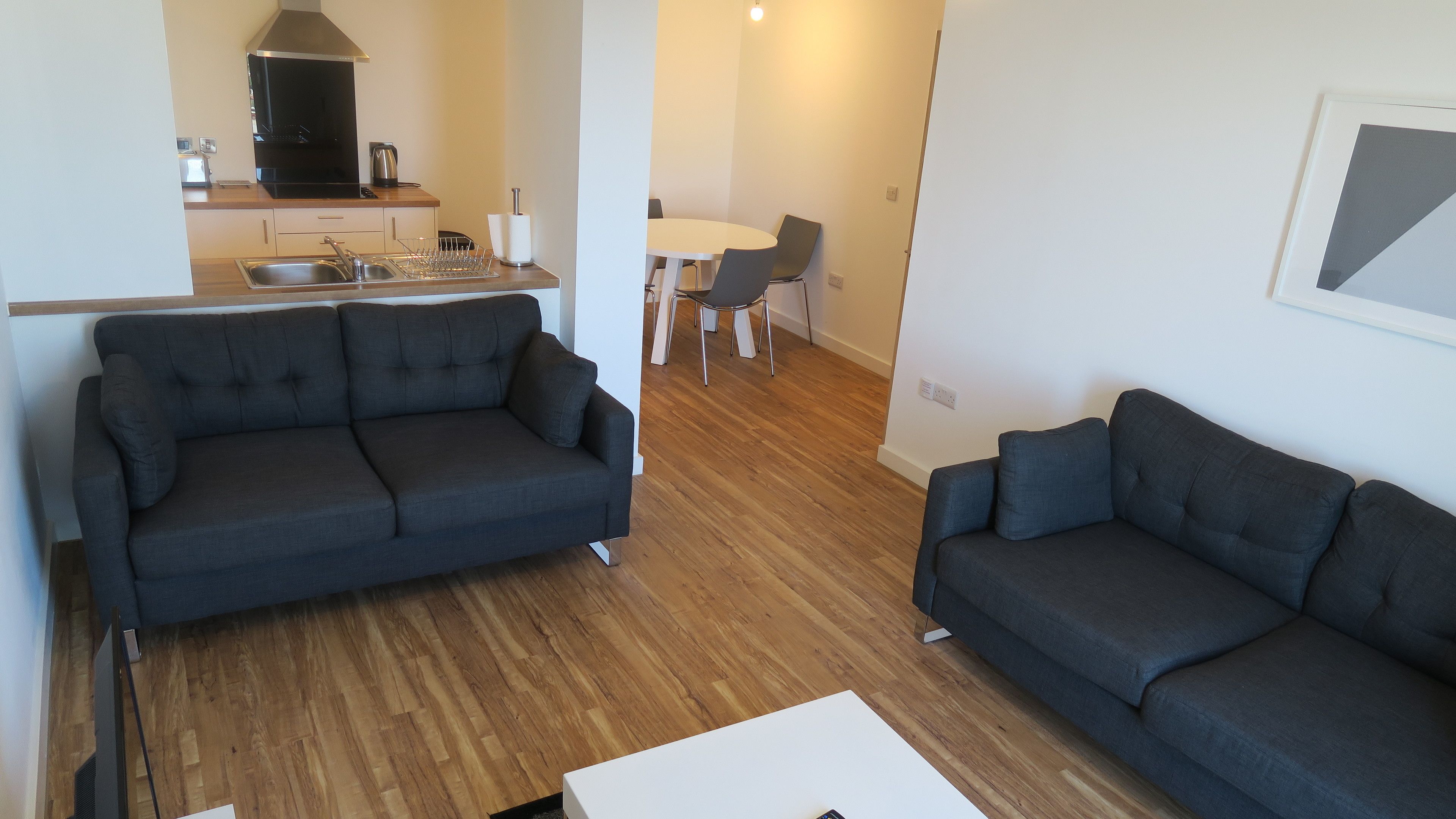 Una sala de estar moderna con dos sofás oscuros, una mesa de centro blanca y una cocina con microondas, hervidor y lavamanos.