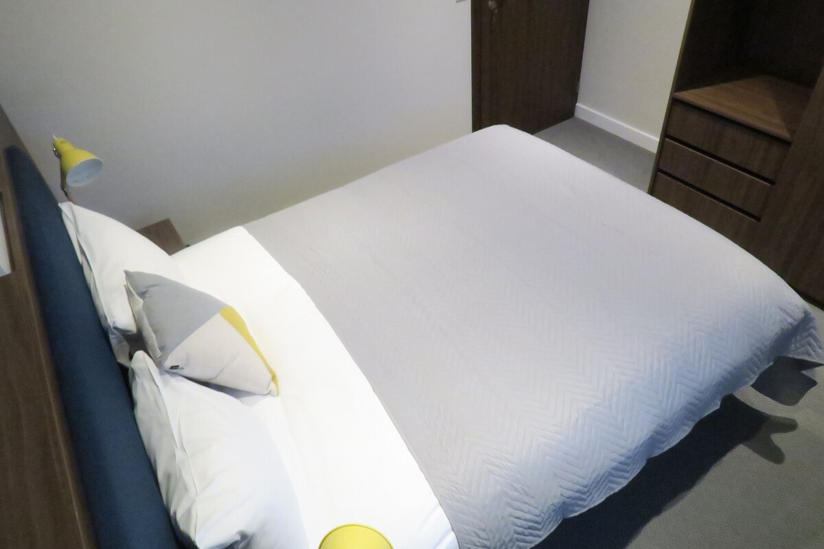 A neatly made bed with white and light blue bedding, a wooden headboard, and a small bedside table with a yellow lamp.