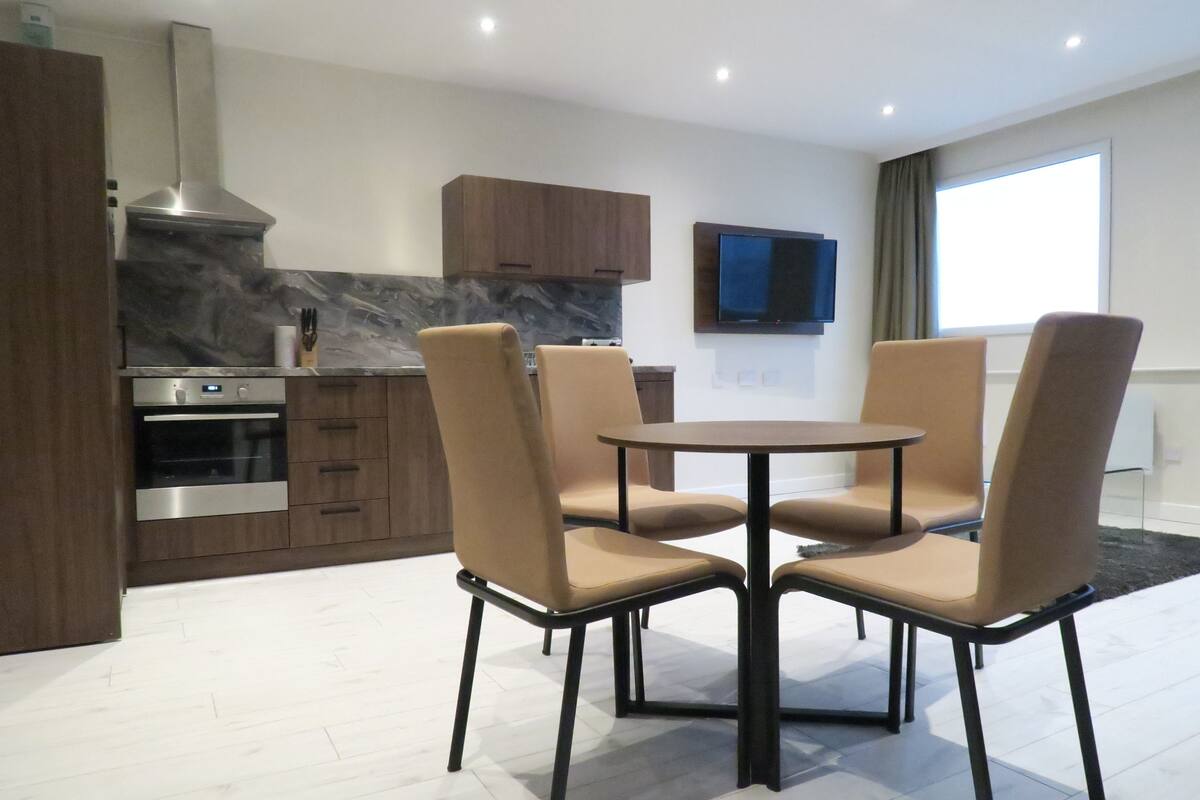 A modern kitchen with a built-in oven, a stainless steel range hood, and wooden cabinets. There is a round dining table with four beige chairs. A flat-screen TV is mounted on the wall above the table.