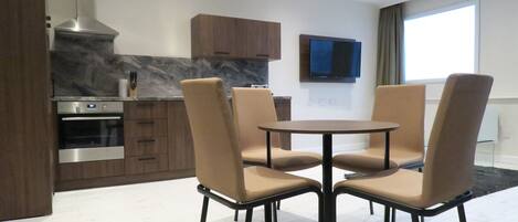 A modern kitchen with a built-in oven, a stainless steel range hood, and wooden cabinets. There is a round dining table with four beige chairs. A flat-screen TV is mounted on the wall above the table.