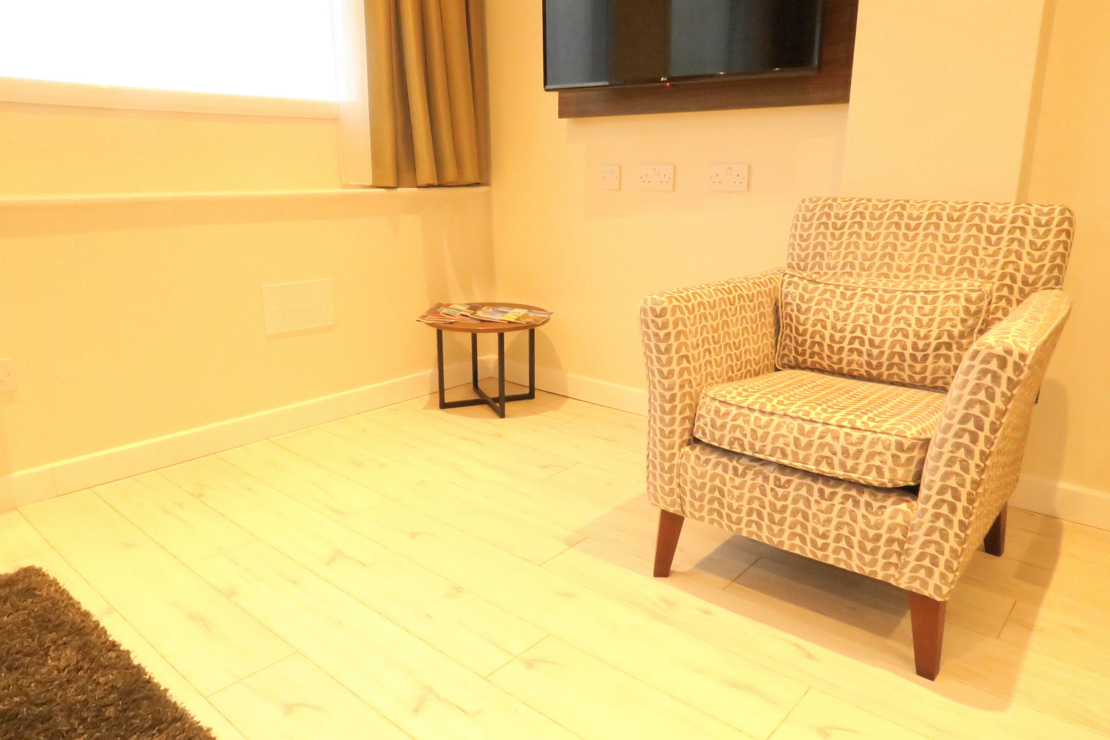 A room with a patterned armchair, a small side table with magazines, a flat-screen TV mounted on the wall, and a window with curtains.