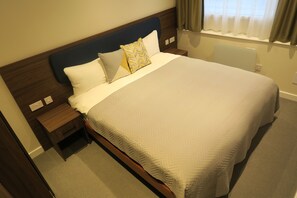 Uma cama bem arrumada com colcha branca e dois travesseiros, cabeceira de madeira, mesa de cabeceira com abajur e janela com cortinas.