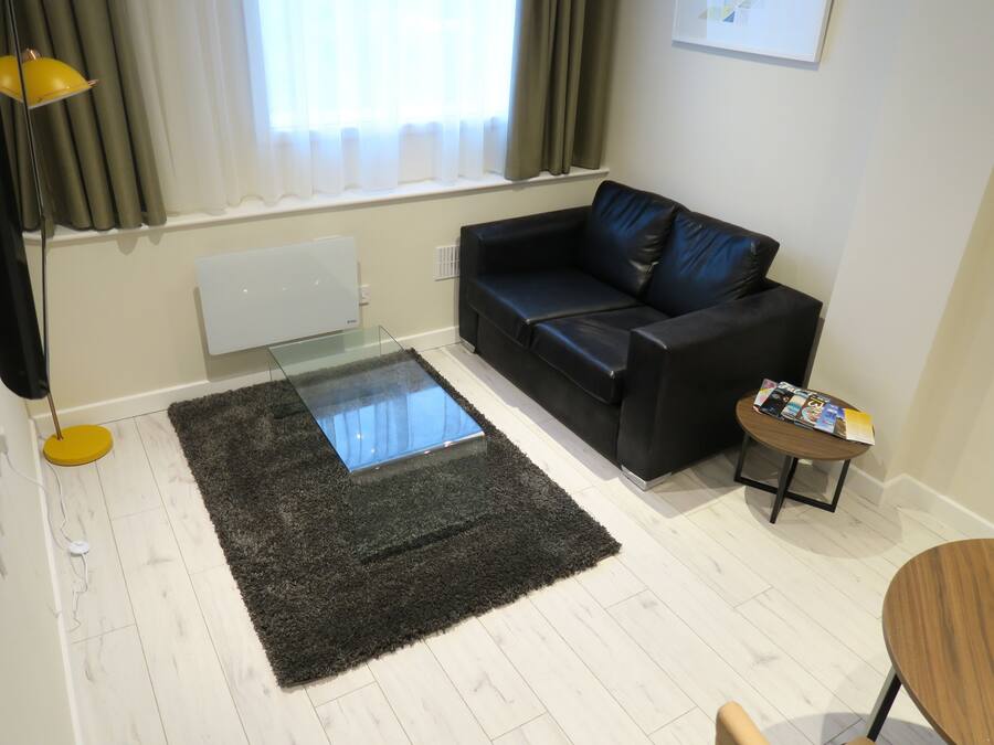 Un salón con un sofá de cuero negro, una mesa de centro de vidrio, una mesita auxiliar de madera y una lámpara de pie amarilla.