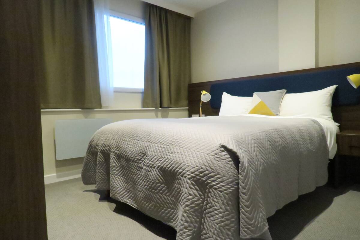 A hotel room with a large bed, a nightstand, a lamp, and a window with curtains.