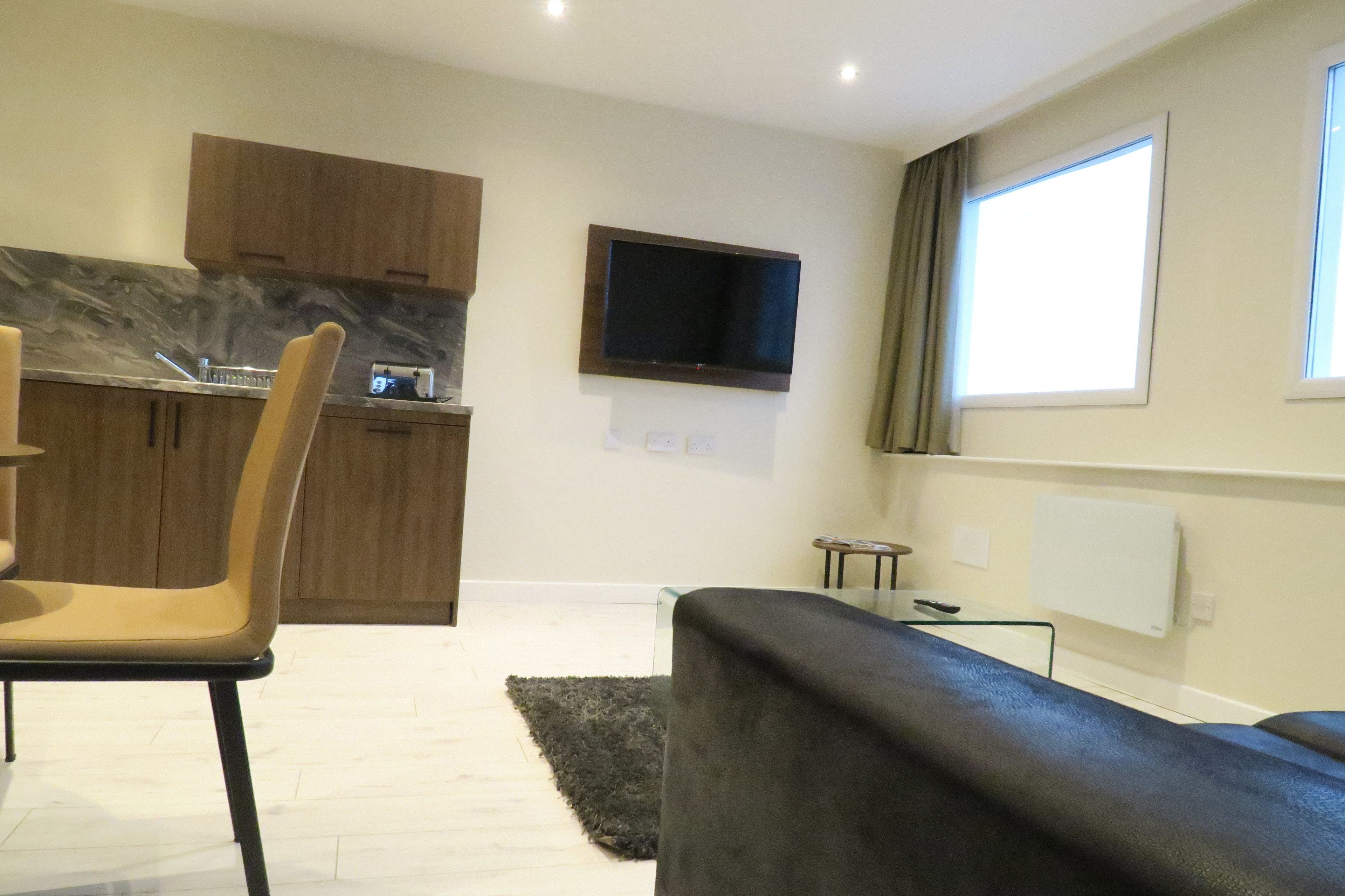A modern living room with a black sofa, a dining area with a chair, a kitchen with wooden cabinets, and a flat-screen TV mounted on the wall.