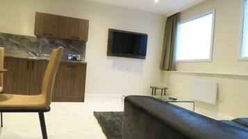 A modern living room with a black sofa, a dining area with a chair, a kitchen with wooden cabinets, and a flat-screen TV mounted on the wall.