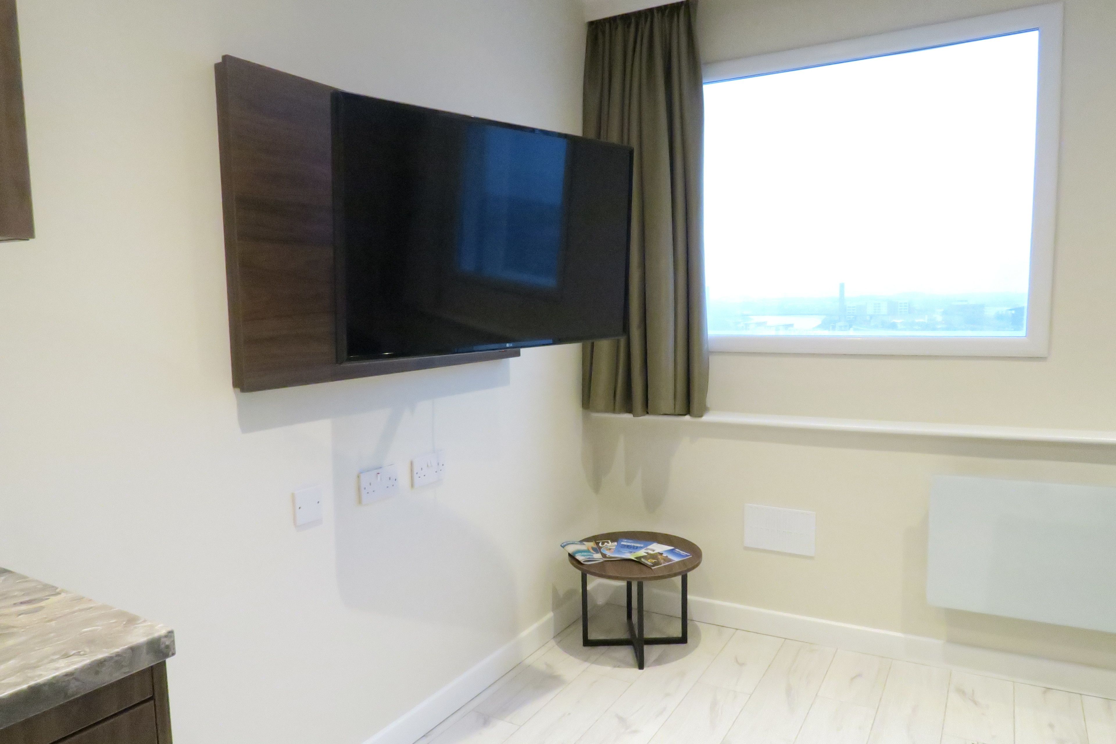 A room with a flat-screen TV mounted on the wall, a small table with magazines, a window with curtains, and a radiator.