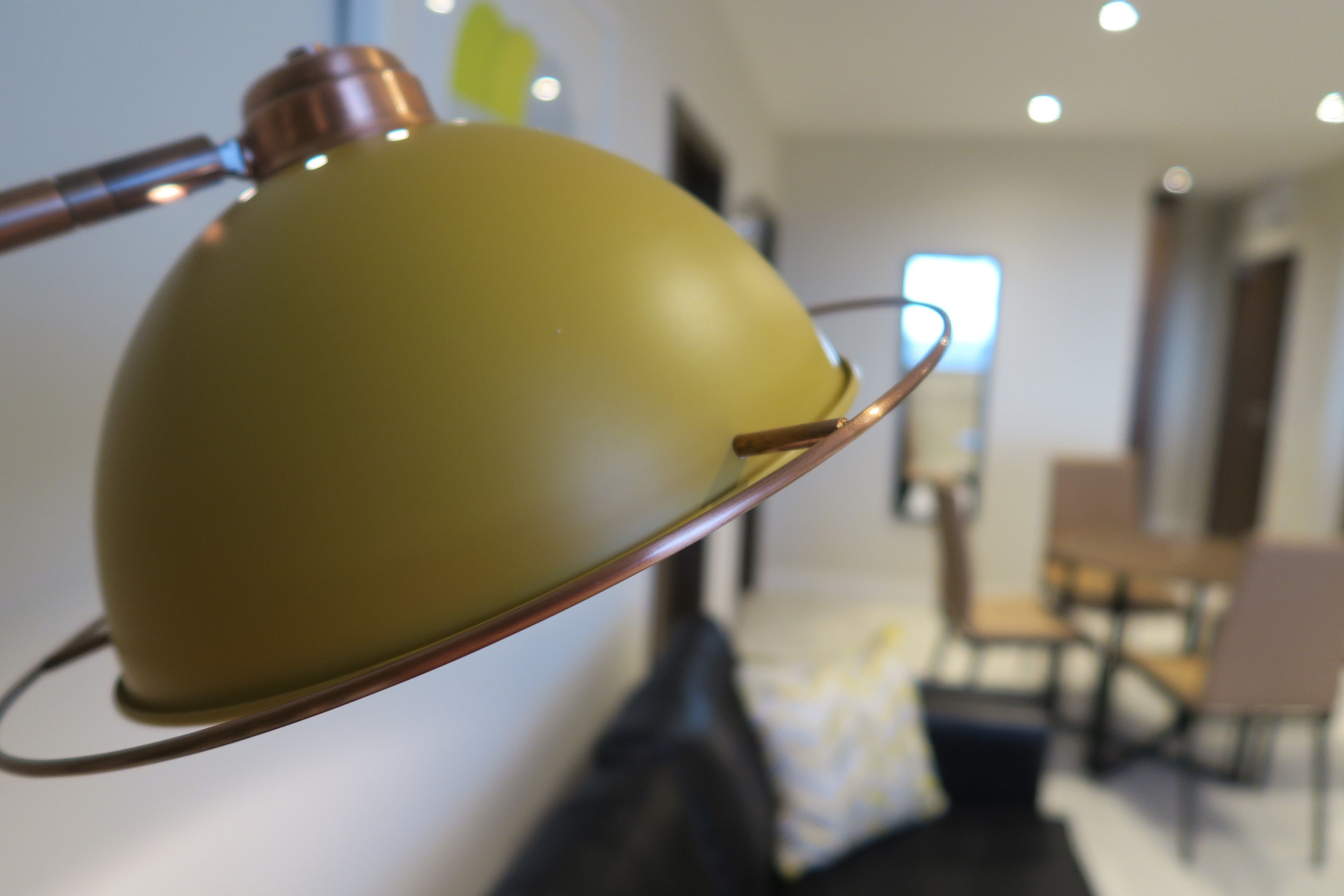 Una lámpara moderna con base metálica y pantalla verde, ubicada en una habitación bien iluminada con un comedor al fondo.